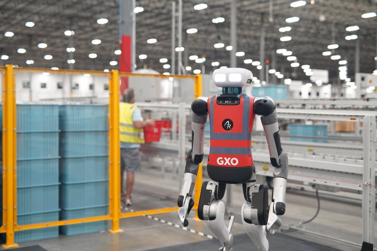 Robot humanoïde Agility Robotics Digit portant une chasuble marquée GXO dans un entrepôt industriel avec convoyeurs et barrières jaunes