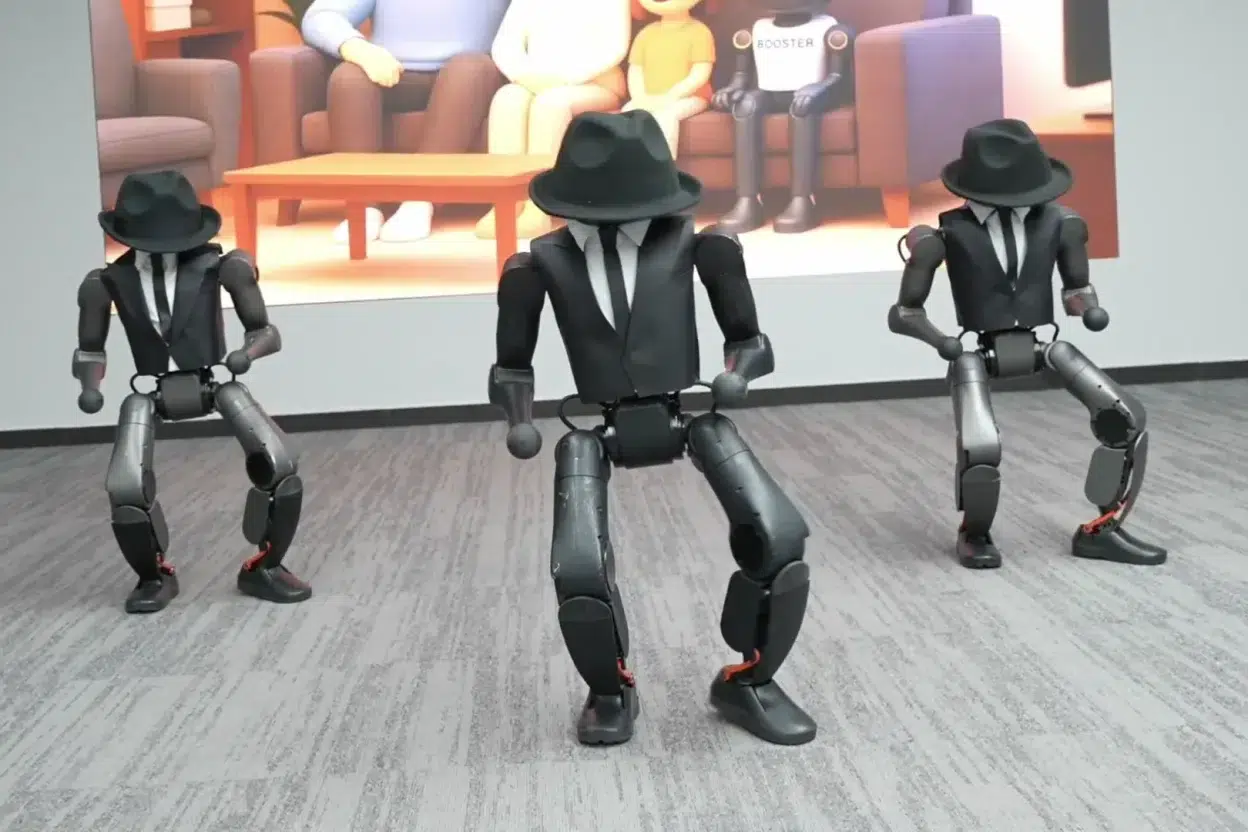 Trois robots humanoïdes Booster Robotics K1 vêtus de costumes et chapeaux noirs, en démonstration devant un écran affichant la marque BOOSTER