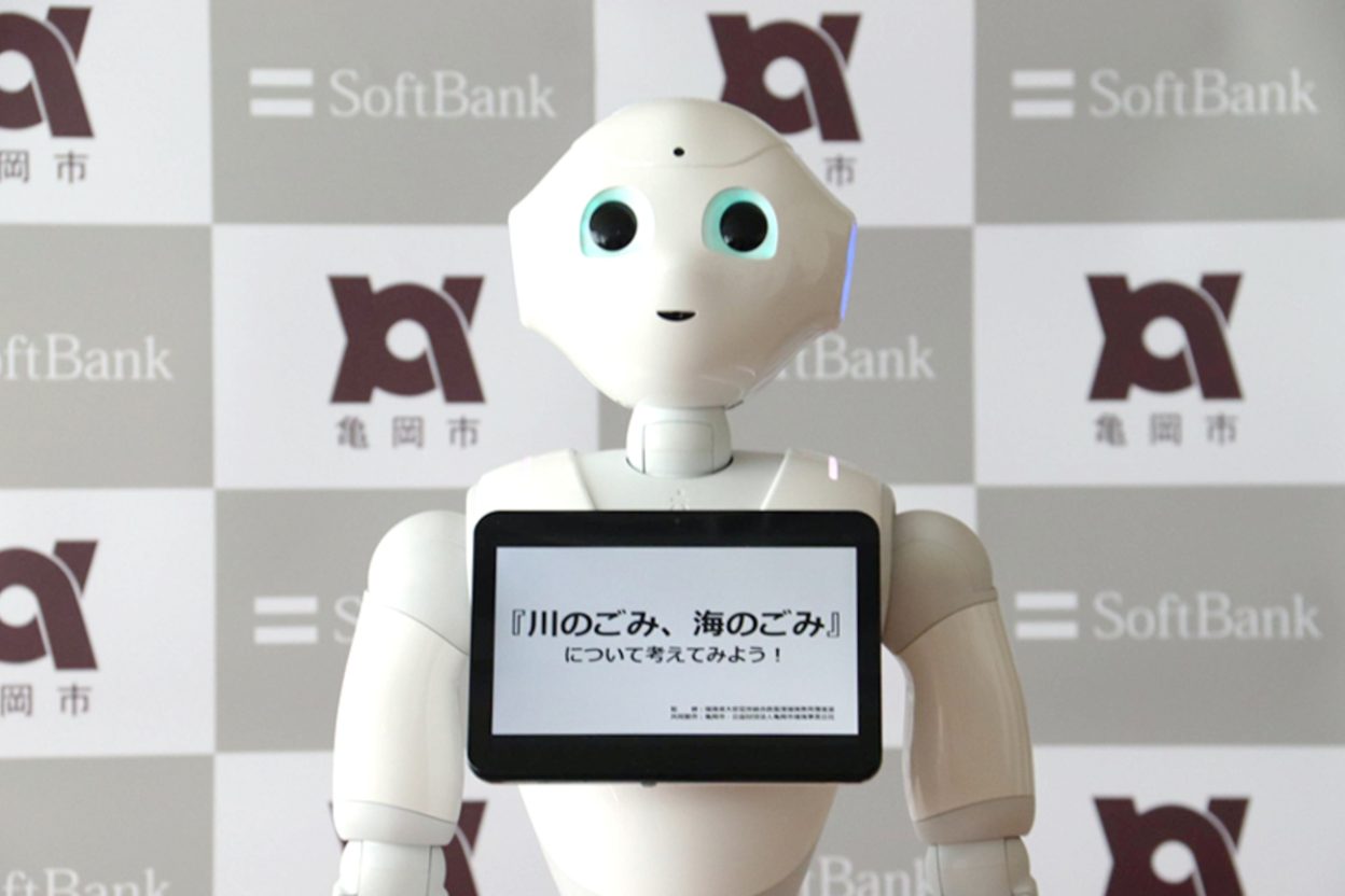 Robot humanoïde SoftBank Robotics Pepper avec tablette tactile intégrée affichant un message en japonais, devant un mur d'écrans SoftBank